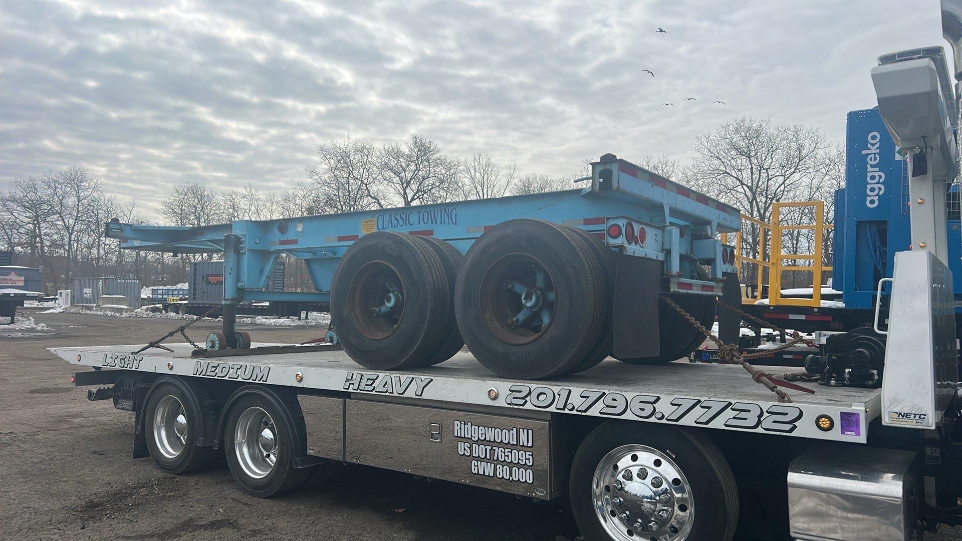 Light blue 20 foot chassis trailer with Classic Towing lettering on the frame, chained down on a flatbed truck in a yard