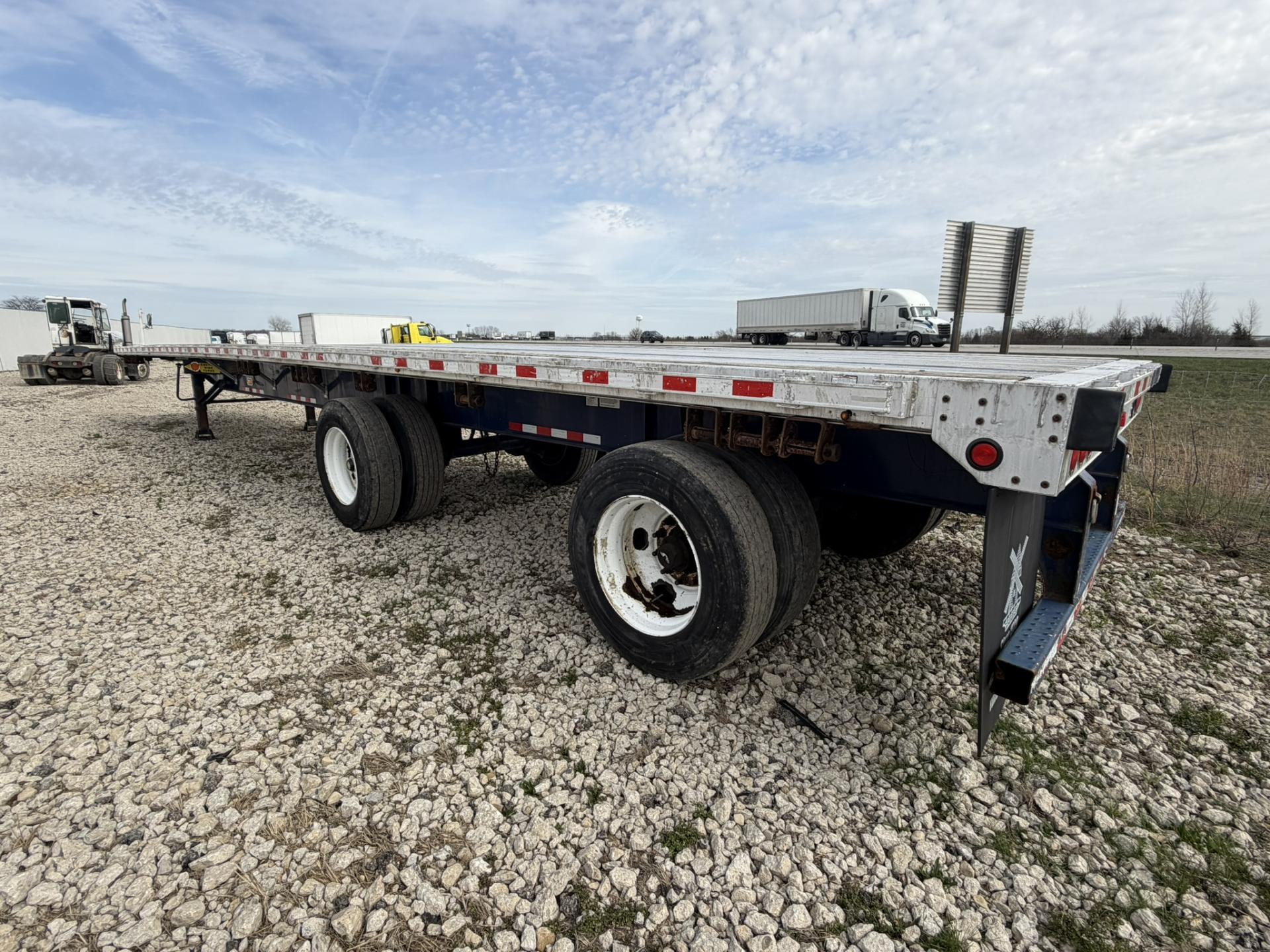 Silver high flatbed trailer with a full-length deck and red-and-white reflective tape, parked in a gravel lot