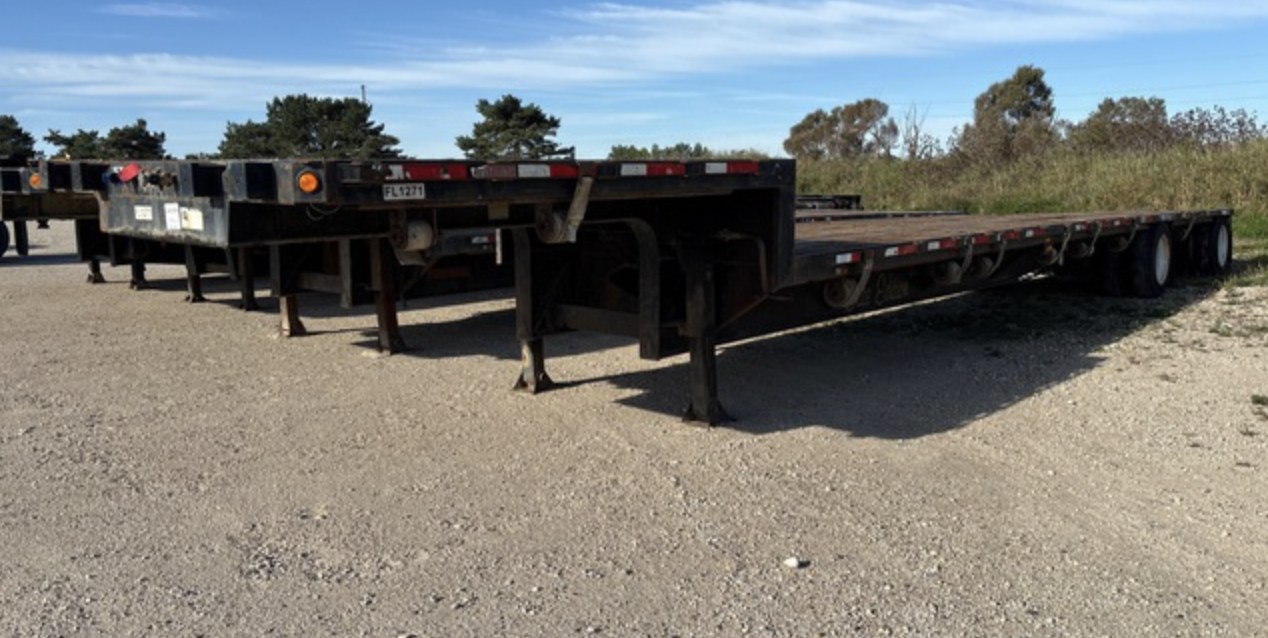 Black step deck trailer with a weathered wooden deck parked in a gravel lot, rear deck dropped below the front