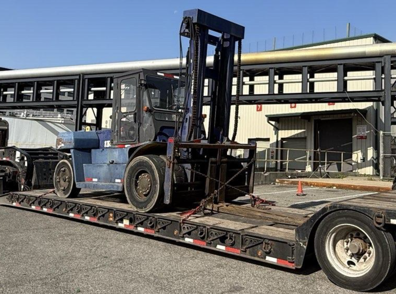Dark blue Komatsu heavy-capacity forklift secured with chains on a flatbed trailer in an industrial yard